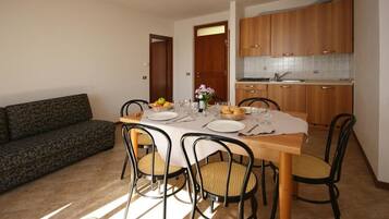 Zona pranzo con tavolo in legno apparecchiato per quattro persone, circondato da sedie in vimini. Sullo sfondo si vede un divano, e una cucina con pensili in legno è adiacente.