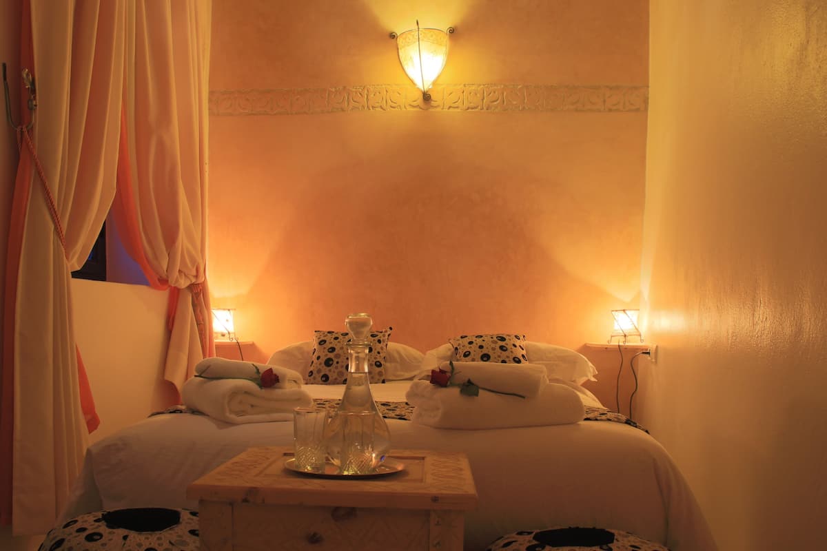 A hotel room with a bed, a bedside table with a bottle and glasses, and two wall-mounted lamps.