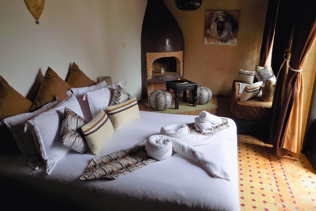 A neatly made bed with multiple pillows and a blanket, a small table with a vase, and a chair with a cushion.