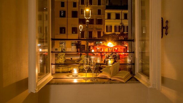 A window with a view of a cityscape at night, featuring buildings and a lit-up bar area. - Palazzo De Cupis (Rome)