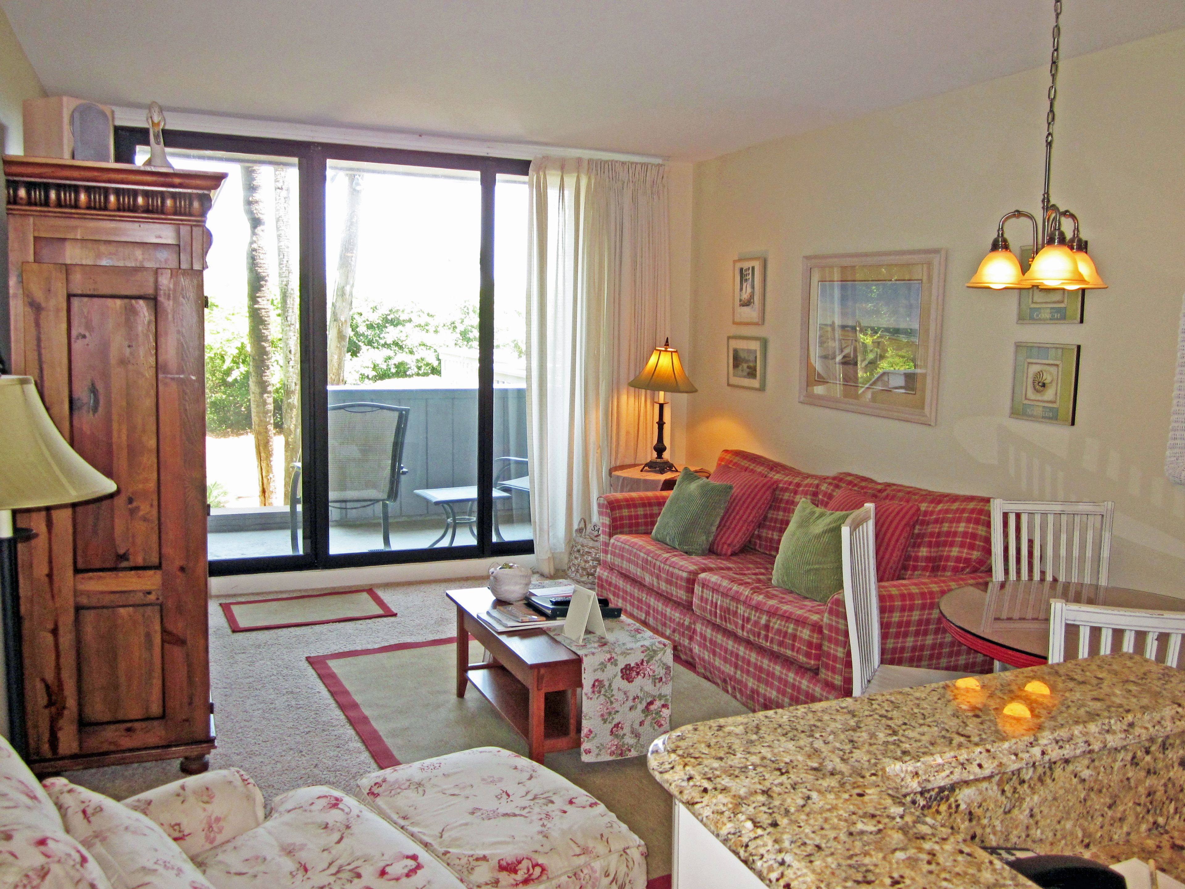 Ein Wohnzimmer mit einem rot-weiß karierten Sofa, einem Holzkabinett, einem Tisch mit einer Lampe und Blick durch eine Glastür auf einen Außenbereich mit Stühlen.