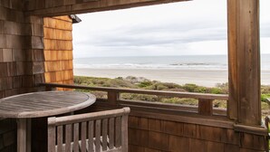 Een houten terras met een tafel en bank, met uitzicht op een strand, de oceaan en begroeiing.