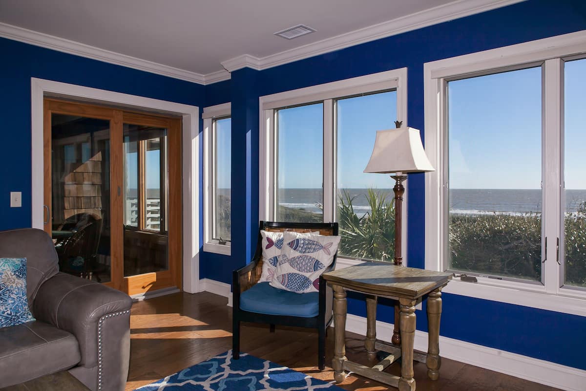 Une pièce avec vue sur l’océan, un mur bleu, une chaise en bois avec un coussin à motifs de poissons, une table avec une lampe et un canapé avec un coussin à motifs.