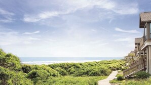 Een pand aan het strand met een houten pad dat leidt naar een gebouw met grote ramen en een balkon met uitzicht op de oceaan.