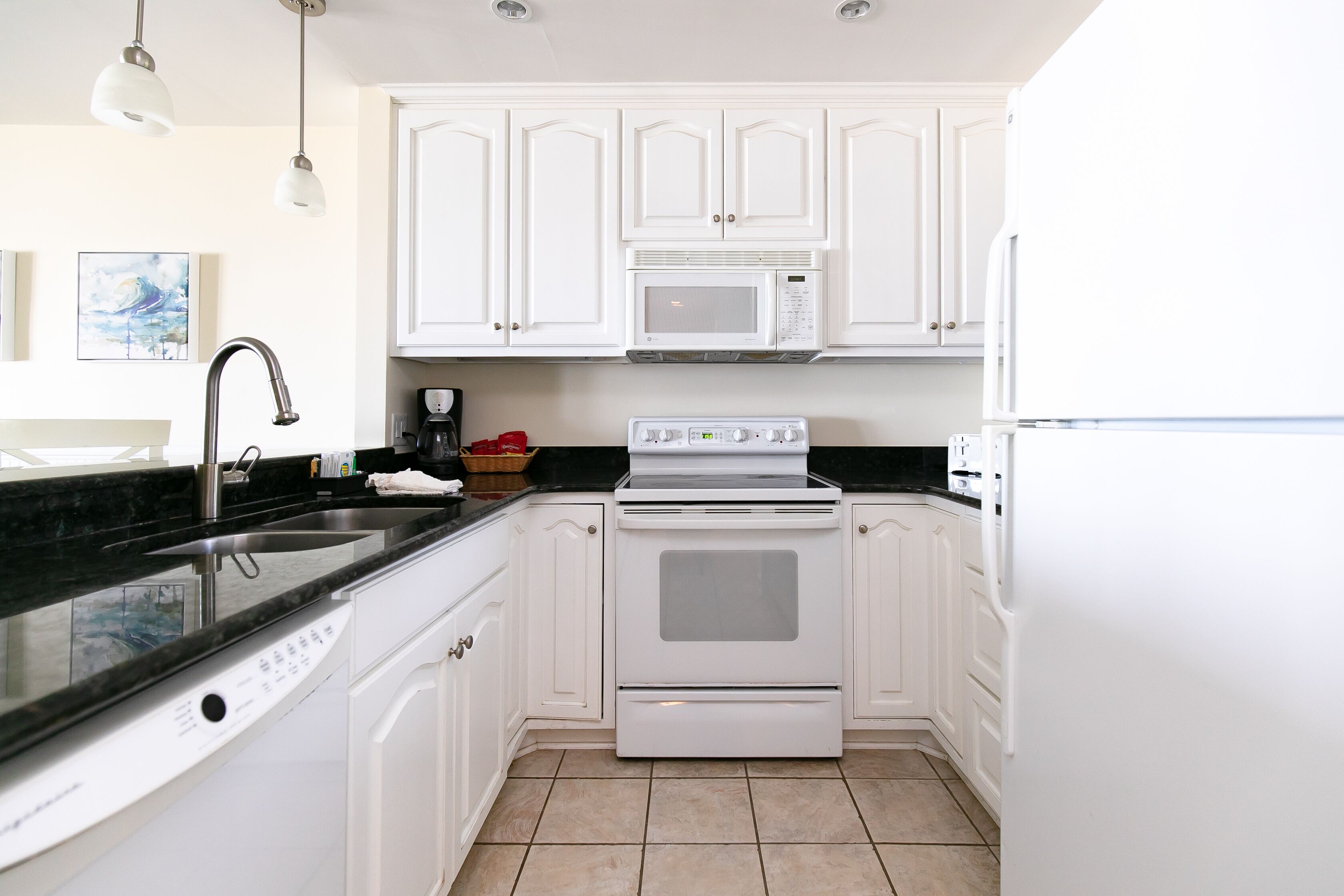 Una cocina con armarios blancos, encimera negra, pileta de acero inoxidable, microondas, horno y refrigerador.