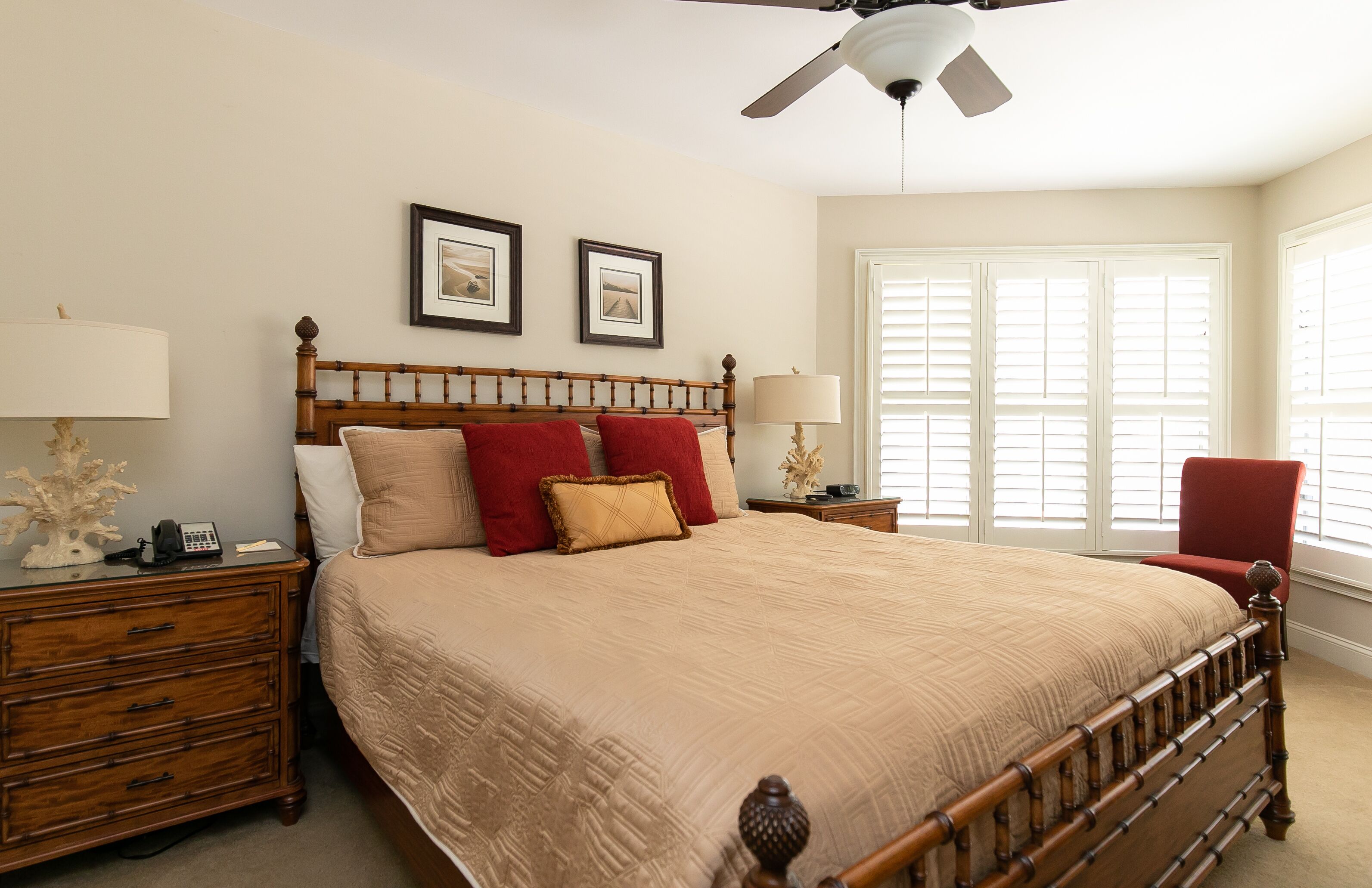 A bedroom with a large bed, wooden headboard, two bedside tables, a nightstand, a red chair, and a ceiling fan.