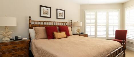 A bedroom with a large bed, wooden headboard, two bedside tables, a nightstand, a red chair, and a ceiling fan.