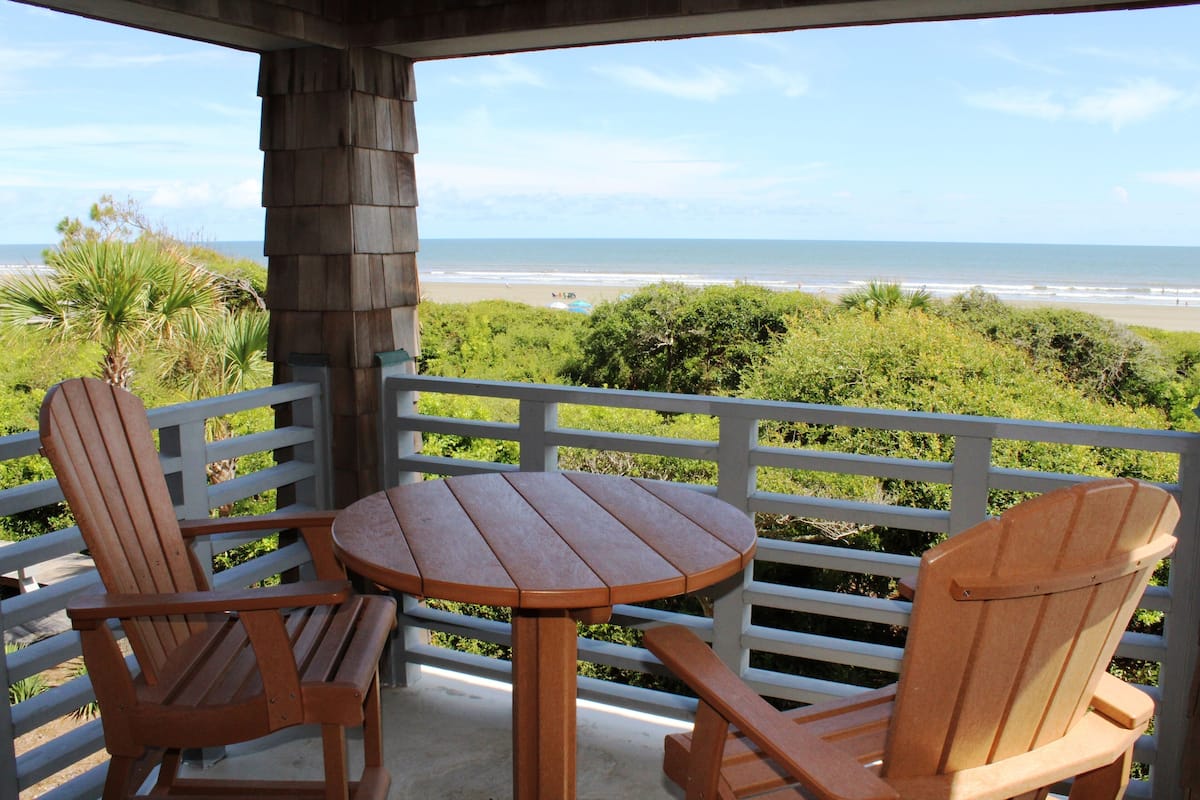 Une terrasse en bois avec deux fauteuils Adirondack et une petite table, donnant sur une plage bordée par l’océan et une végétation luxuriante.