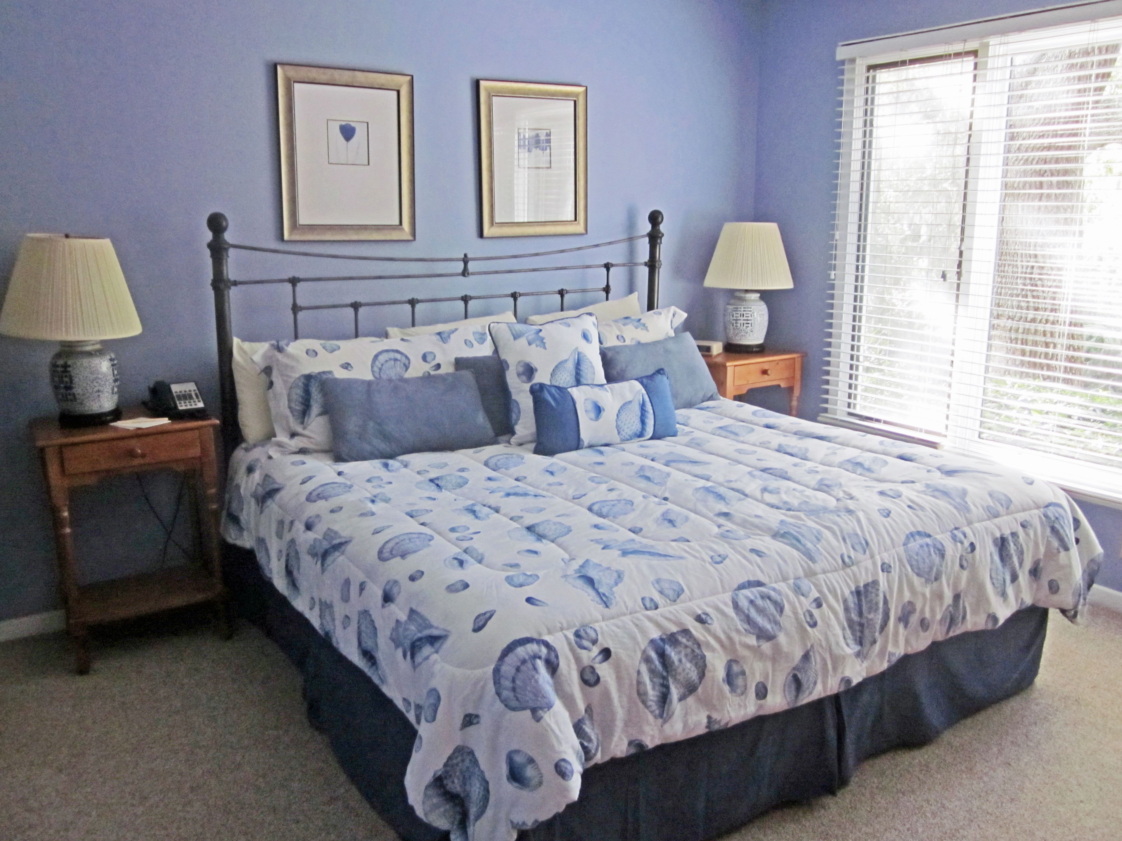 A bedroom with a bed, two bedside tables, two lamps, a window with blinds, and two framed pictures on the wall.