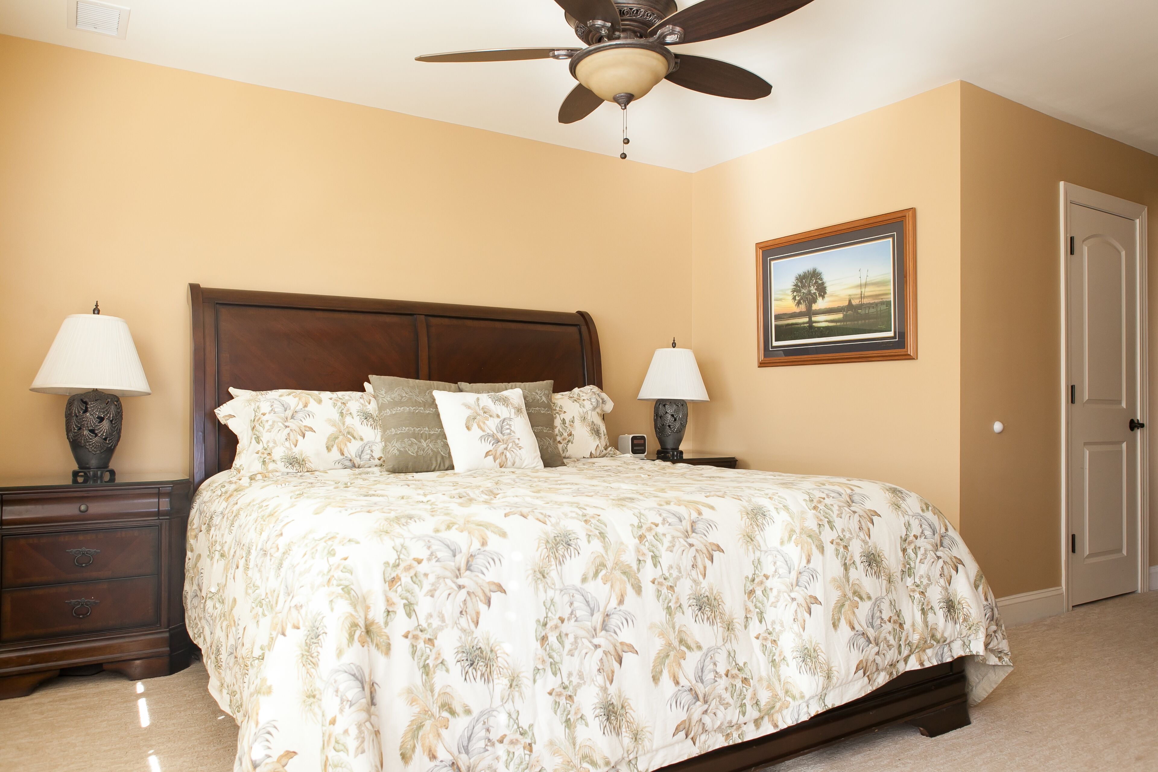 A bedroom with a large bed, two bedside tables with lamps, a ceiling fan, a door, and a framed picture on the wall.