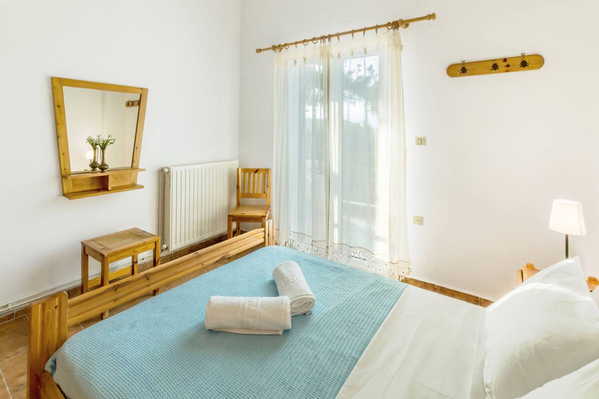 Un dormitorio con cama, una silla de madera, una mesita, un espejo y una ventana con cortinas.