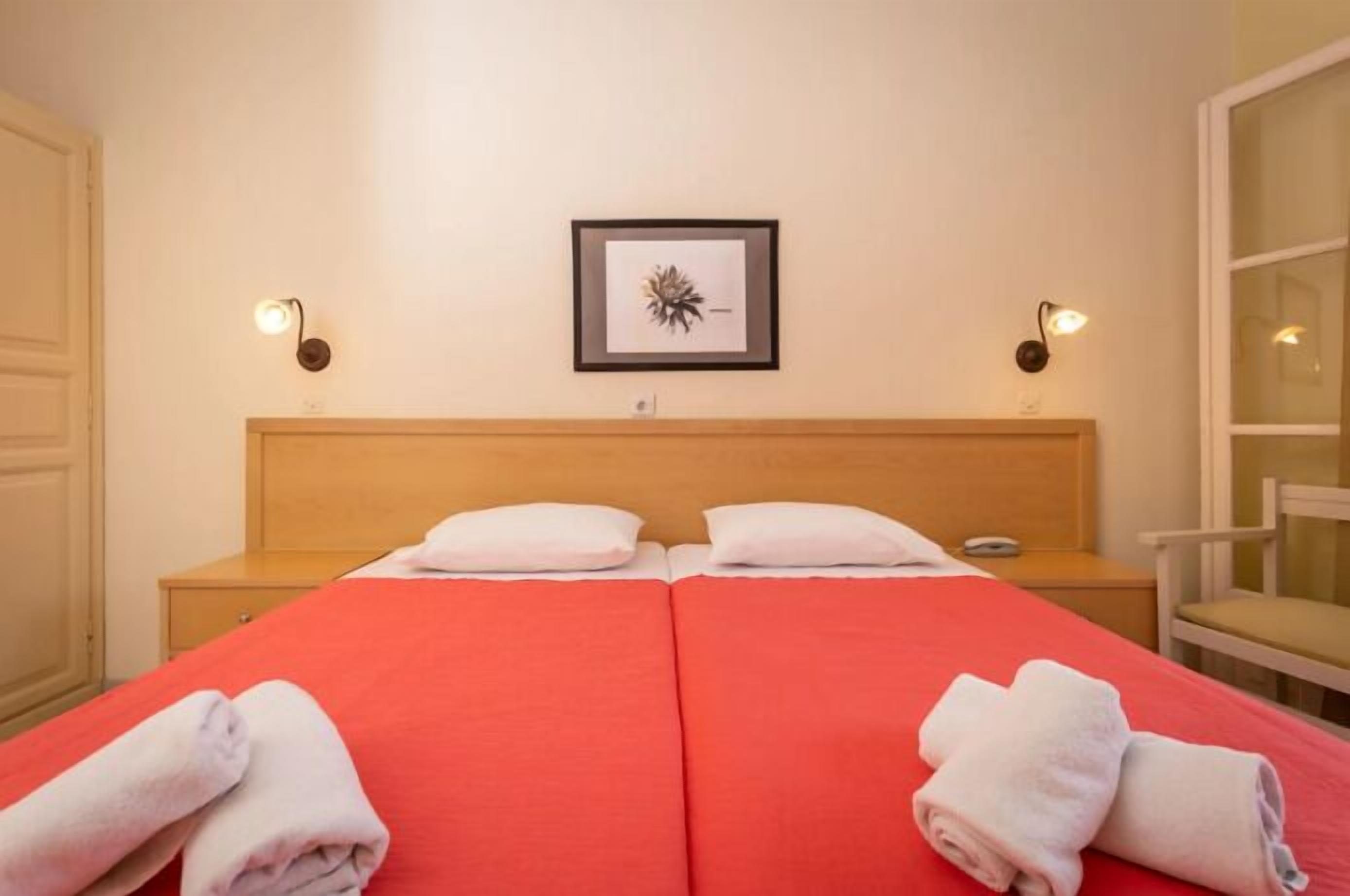A double bed with red bedding, two white pillows, and two rolled towels at the foot of the bed. There is a framed picture on the wall above the bed.