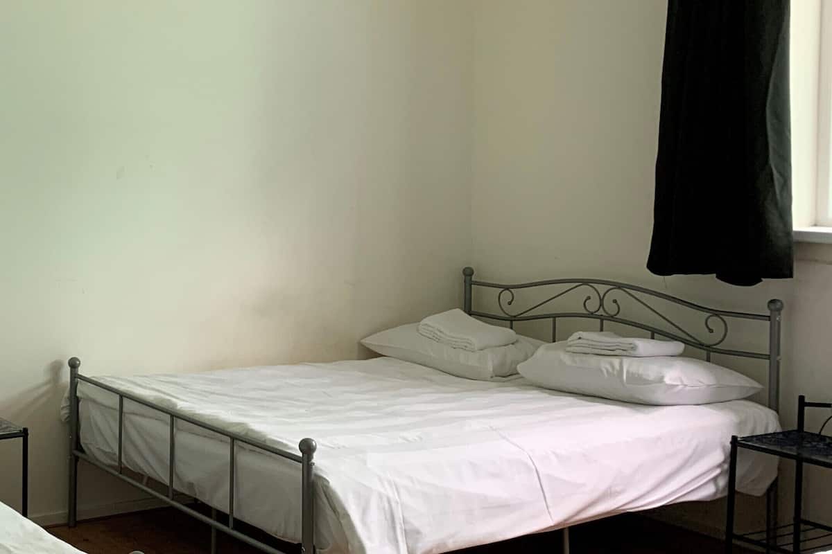 Une petite chambre avec un lit simple, un cadre en métal, du linge de lit blanc et un rideau noir devant la fenêtre.