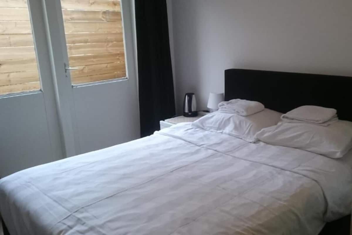 Un lit bien fait, avec du linge de lit blanc, une tête de lit noire, une table de chevet avec une lampe et une fenêtre avec des stores.