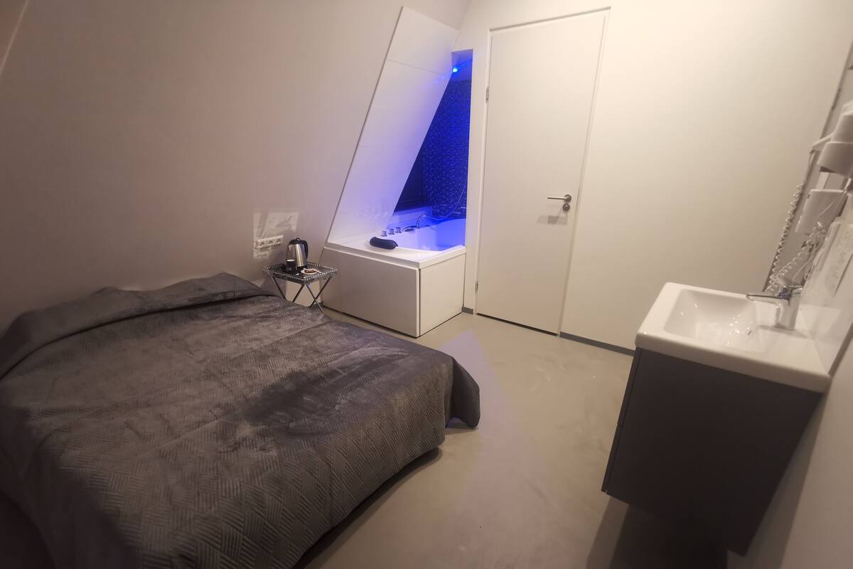 Une chambre à coucher avec un lit, une table de chevet avec une lampe, une petite fenêtre et une salle de bain avec un lavabo et un miroir.