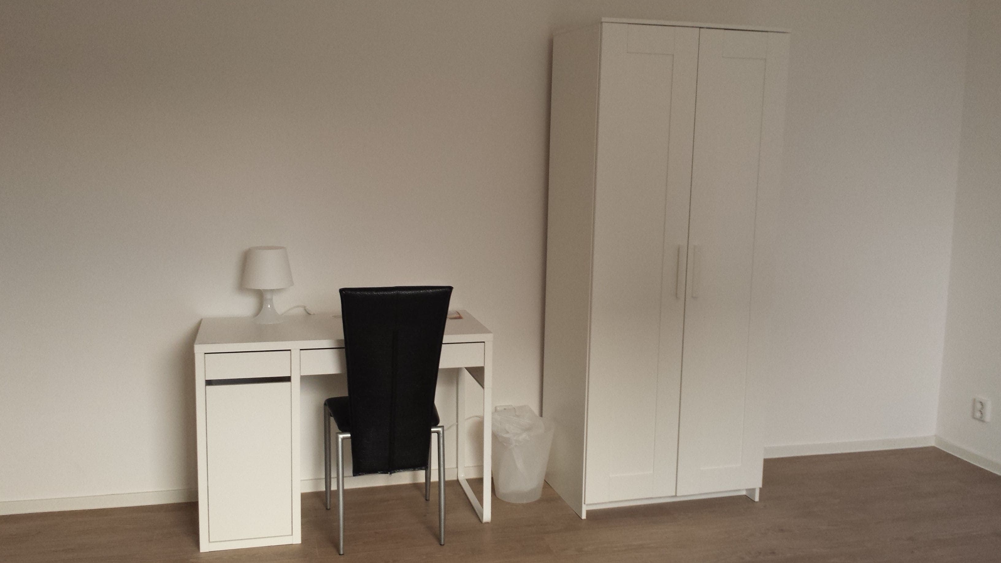 A room with a white desk, a black chair, a white wardrobe, and a white trash bin.