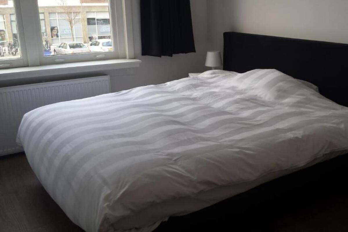 Un lit bien fait, avec une literie blanche, dans une pièce dotée d’une fenêtre, d’un radiateur et d’une table de chevet avec une lampe.