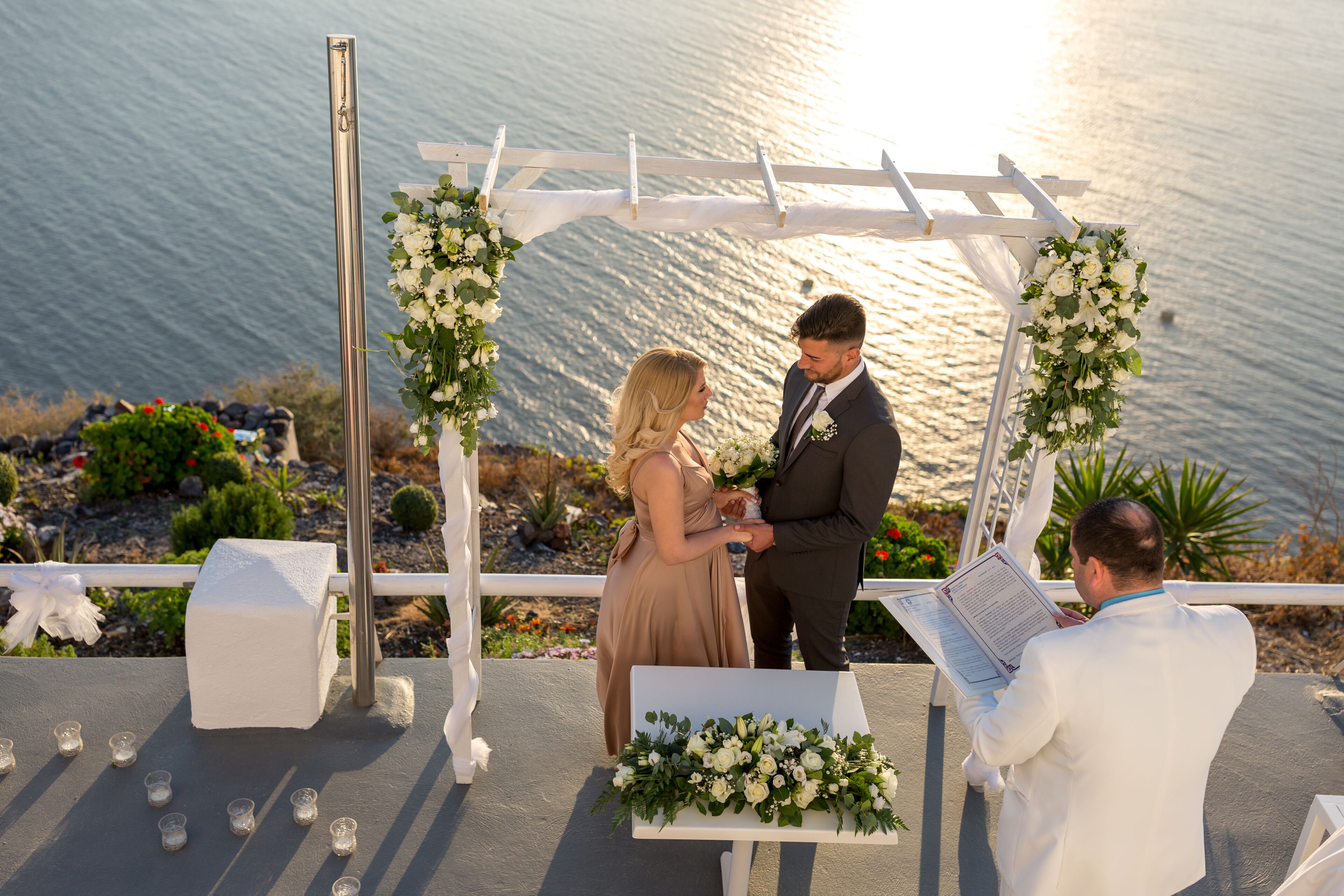 A wedding ceremony with a white arch and floral decorations, set against a scenic backdrop of water and greenery.