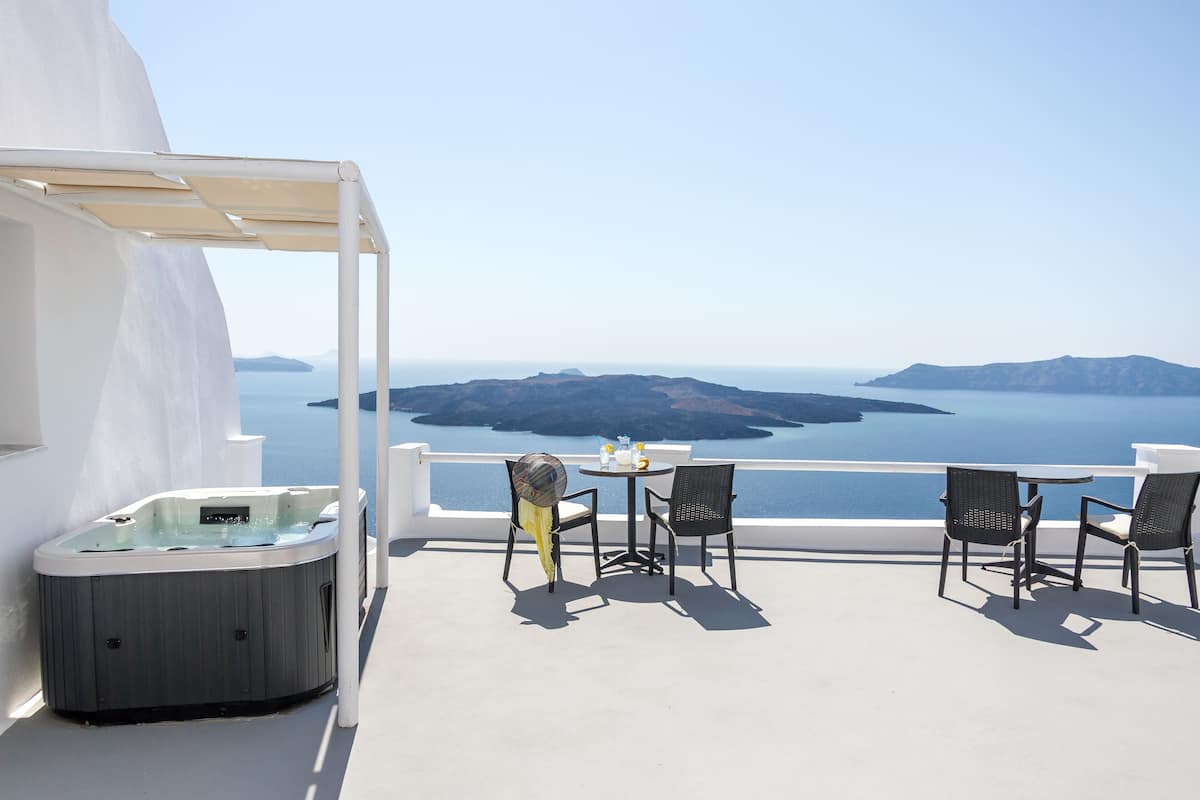 A balcony with a hot tub, two chairs, and a table with bottles, overlooking a scenic view of islands and the sea.