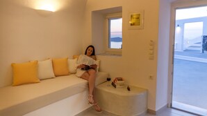 A woman sitting on a white sofa with yellow cushions, reading a book. There is a window with a view of the sea and a door leading outside. - Asma Suites (Santorini)