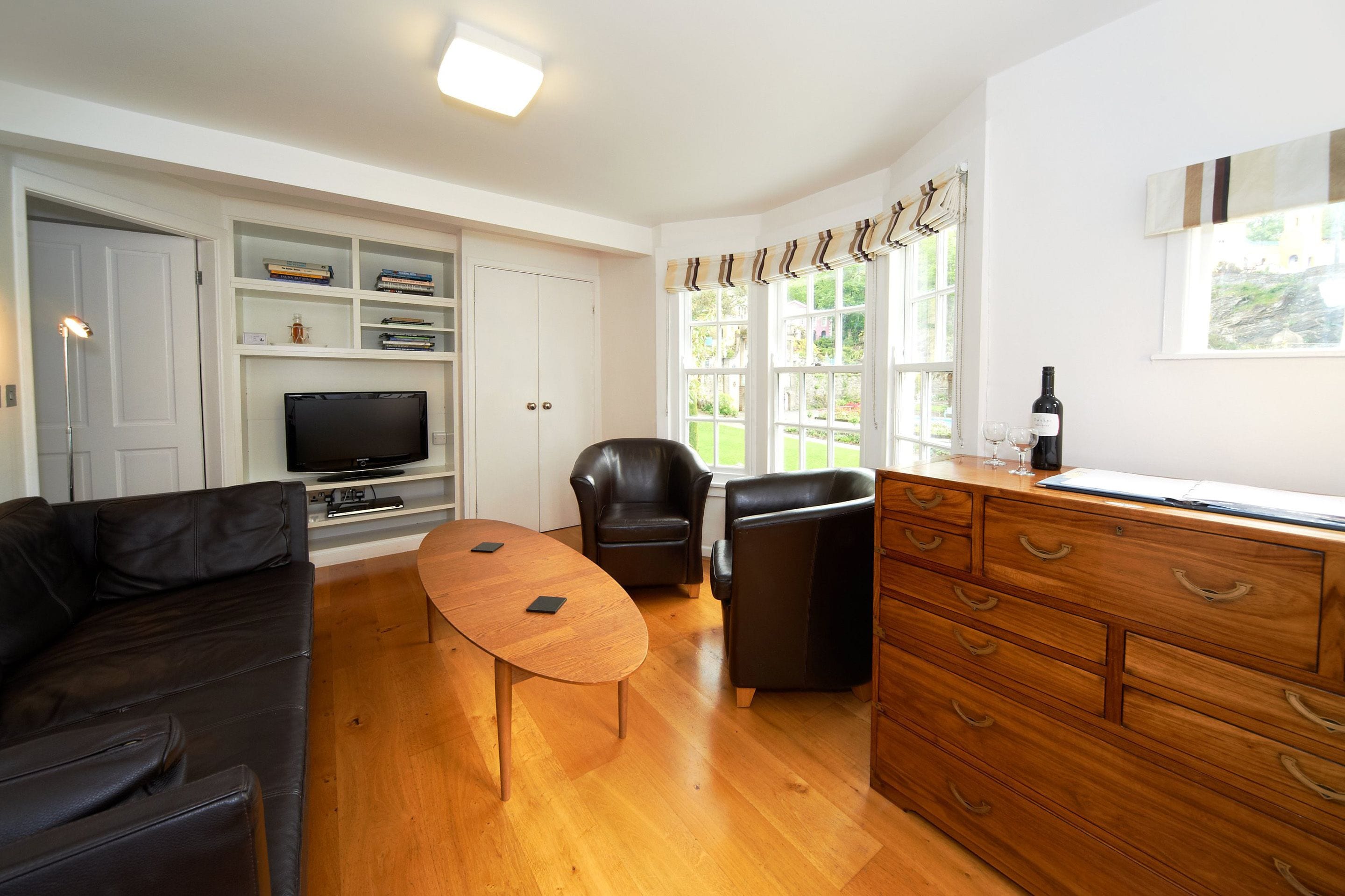 A modern living room with a wooden coffee table, black leather sofas, a TV on a shelf, and a wooden cabinet with drawers.