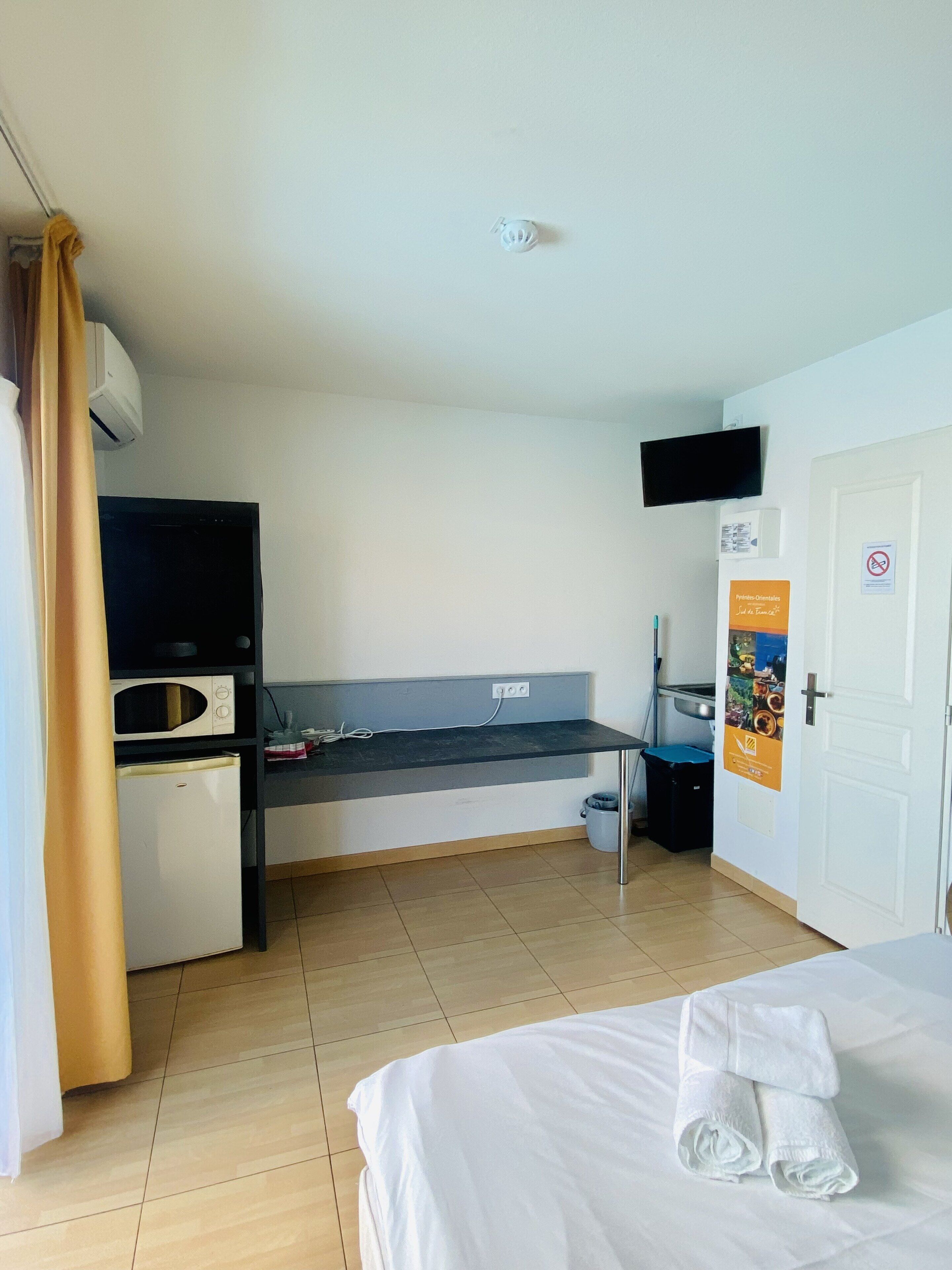 A hotel room with a bed, a small kitchenette, a TV mounted on the wall, and a door with a sign.