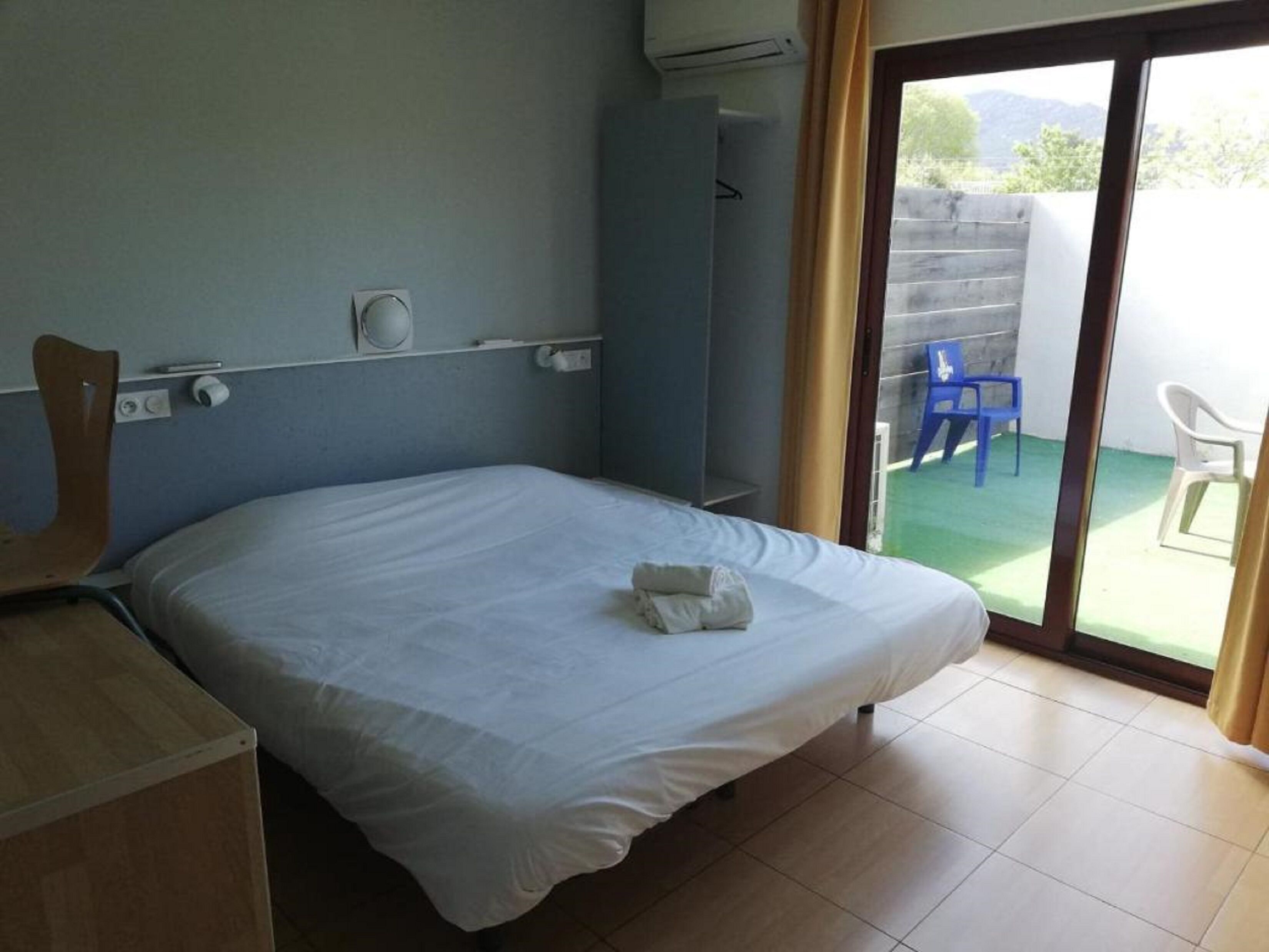 A single-bed room with a wooden headboard, a small desk, and a window with a view of outdoor chairs and greenery.
