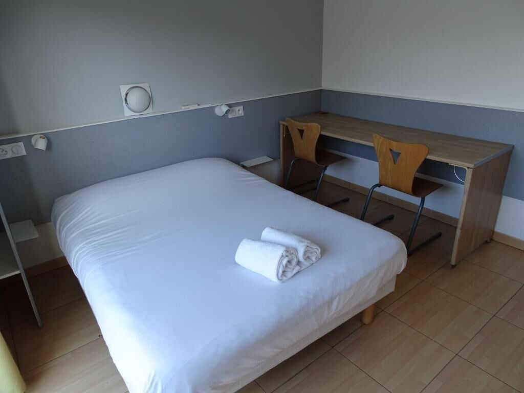 Una cama individual con colchón blanco y una toalla doblada con cuidado, un escritorio de madera con silla, y un enchufe de pared.