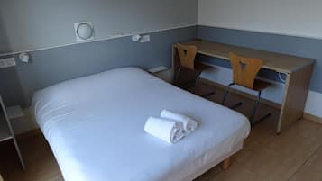 Una cama individual con colchón blanco y una toalla doblada con cuidado, un escritorio de madera con silla, y un enchufe de pared.