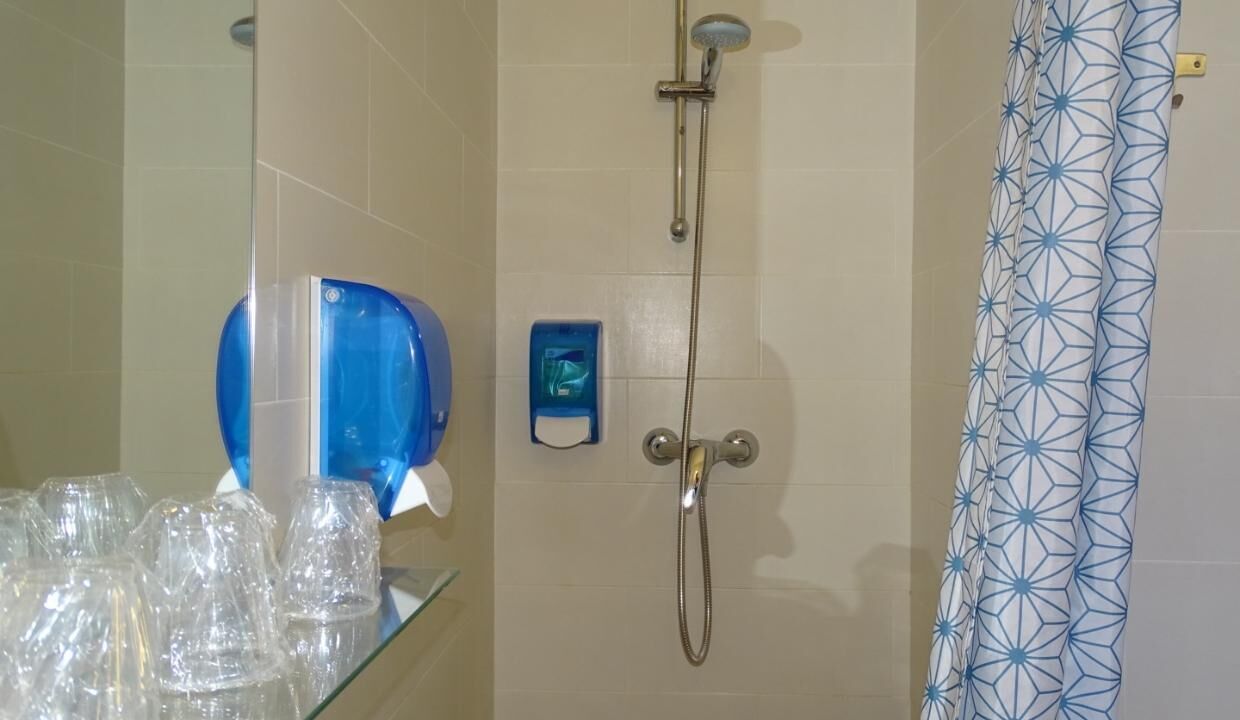 Un baño con ducha, un estante de vidrio con vasos transparentes y un dispensador de jabón azul.