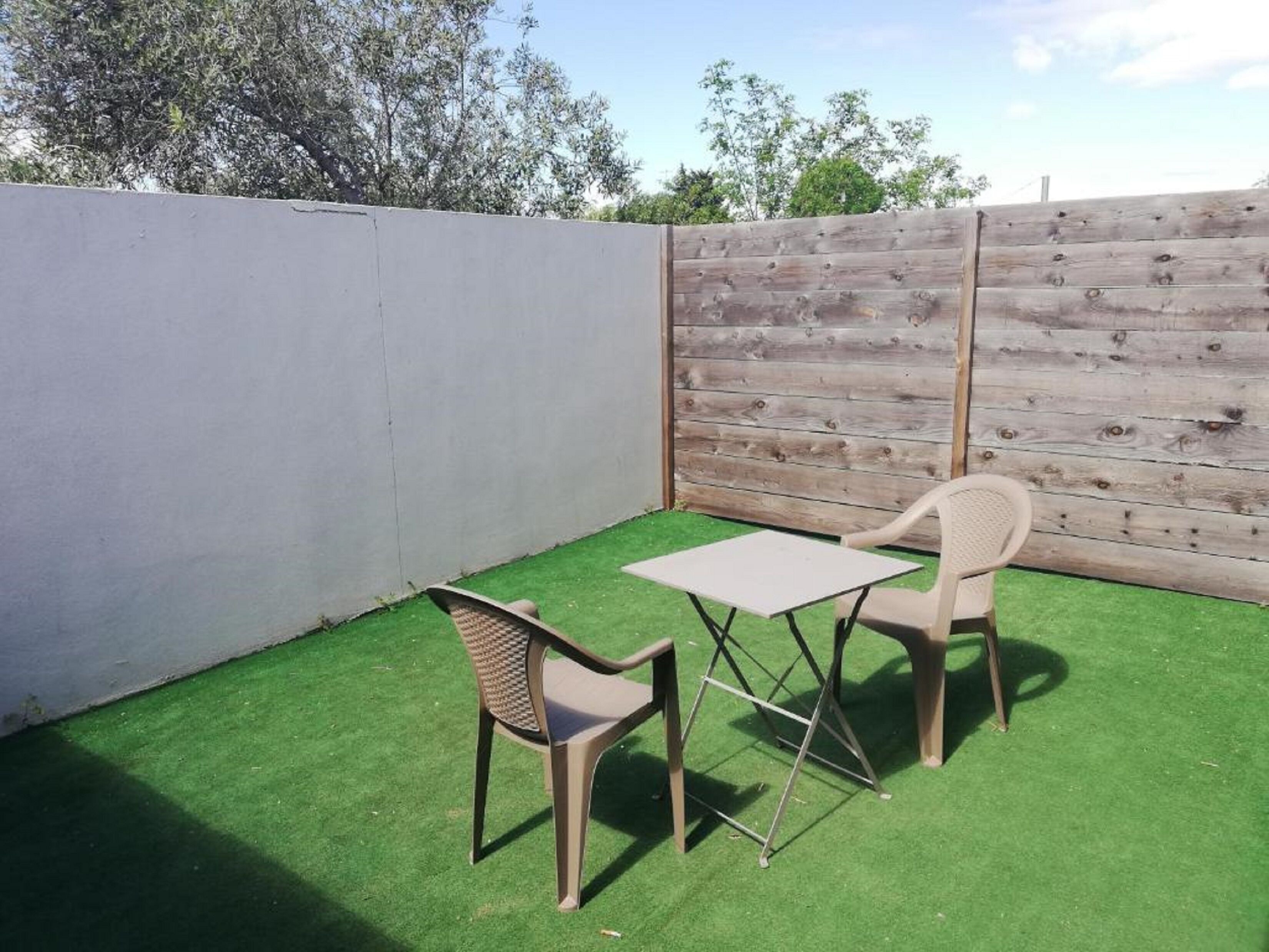 Un petit espace extérieur aménagé en patio, avec du gazon synthétique, deux chaises en plastique et une petite table.