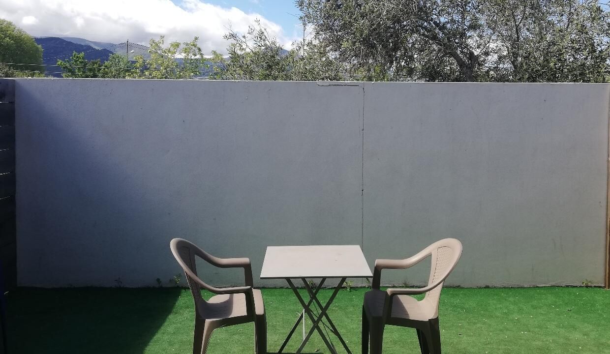 A small outdoor patio with a table and two chairs, a grey wall, and green artificial grass.