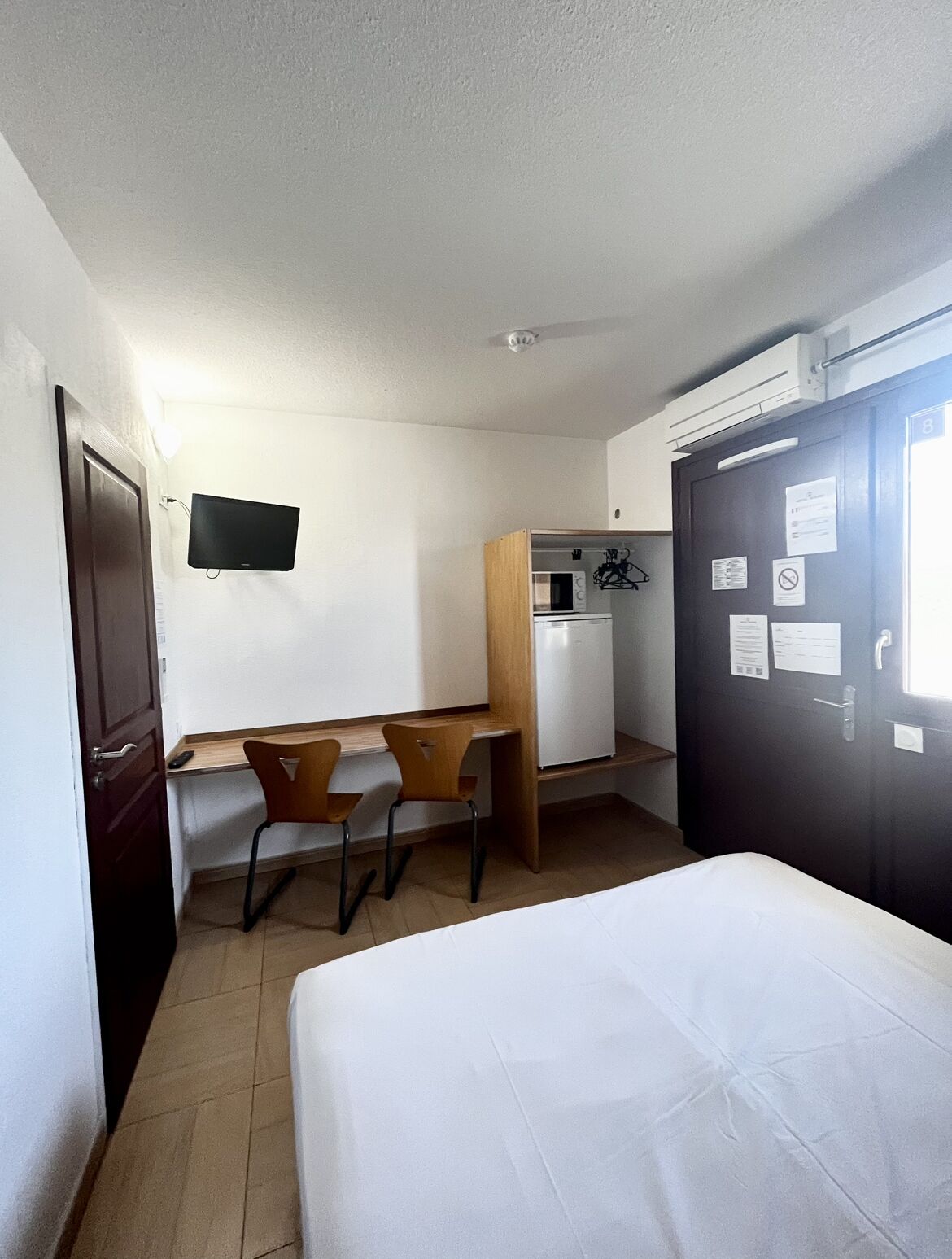 Een kleine hotelkamer met een bed, een bureau met twee stoelen, een kitchenette en een donkere deur met papieren.