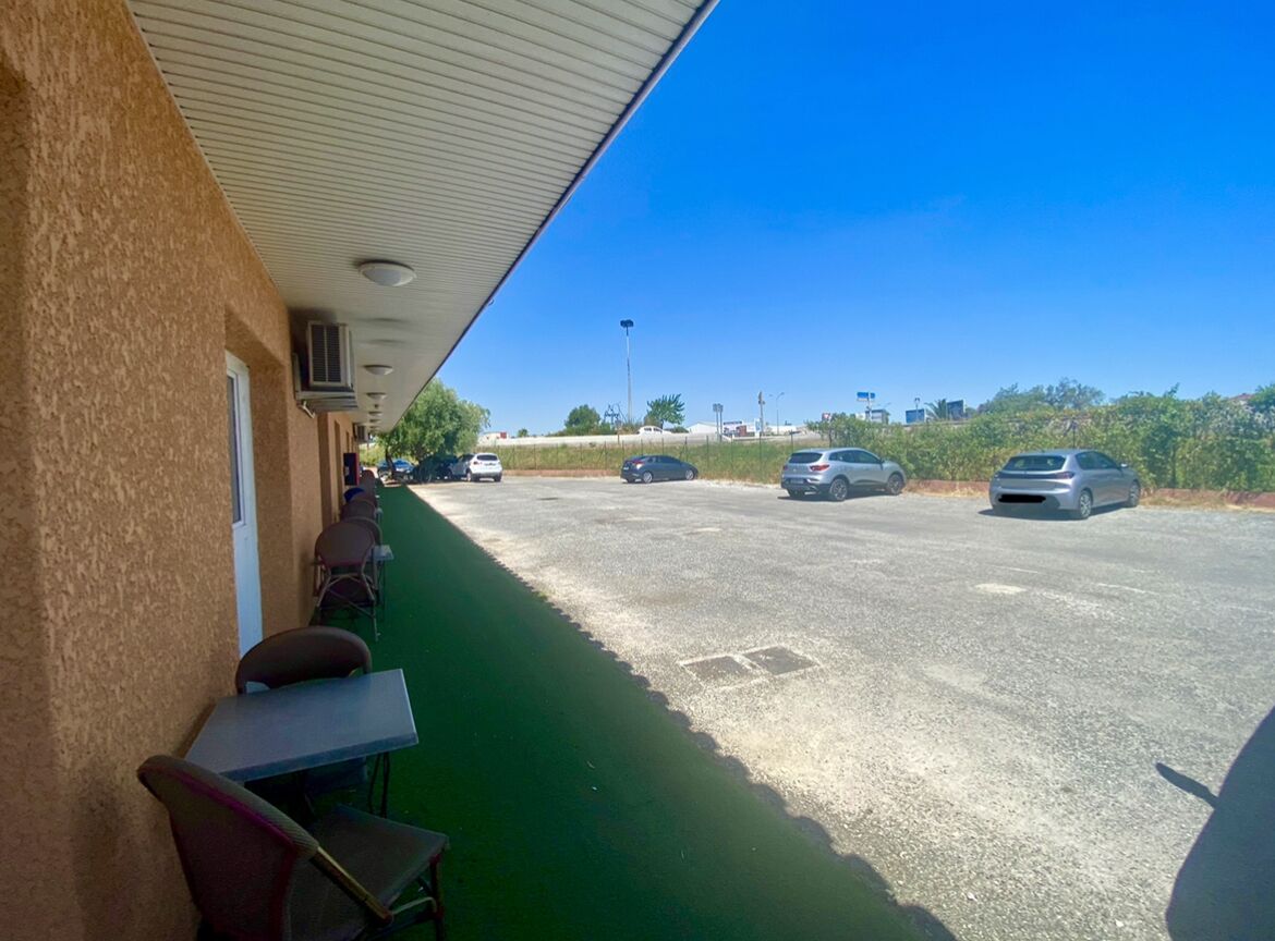 Un espace extérieur abrité, équipé de tables et de chaises, un parking avec plusieurs voitures et un ciel bleu dégagé.