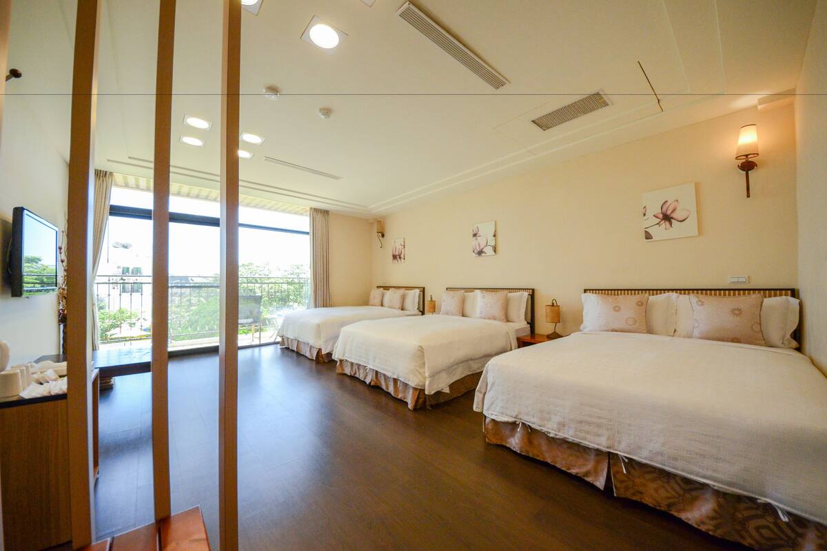 Habitación de hotel con tres camas, televisor y balcón con vistas a zonas verdes.