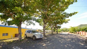 A parked car in a shaded area with trees, a yellow building, and a hilly landscape in the background. - Ciao House (Hengchun)