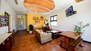 A spacious room with a wooden ceiling, a large woven sofa, a wooden dining table, and a potted plant. - Ciao House (Hengchun)