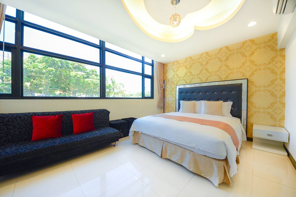 Habitación de hotel con una cama grande, un sofá con cojines rojos, una ventana con vistas a árboles y una pared con diseño.