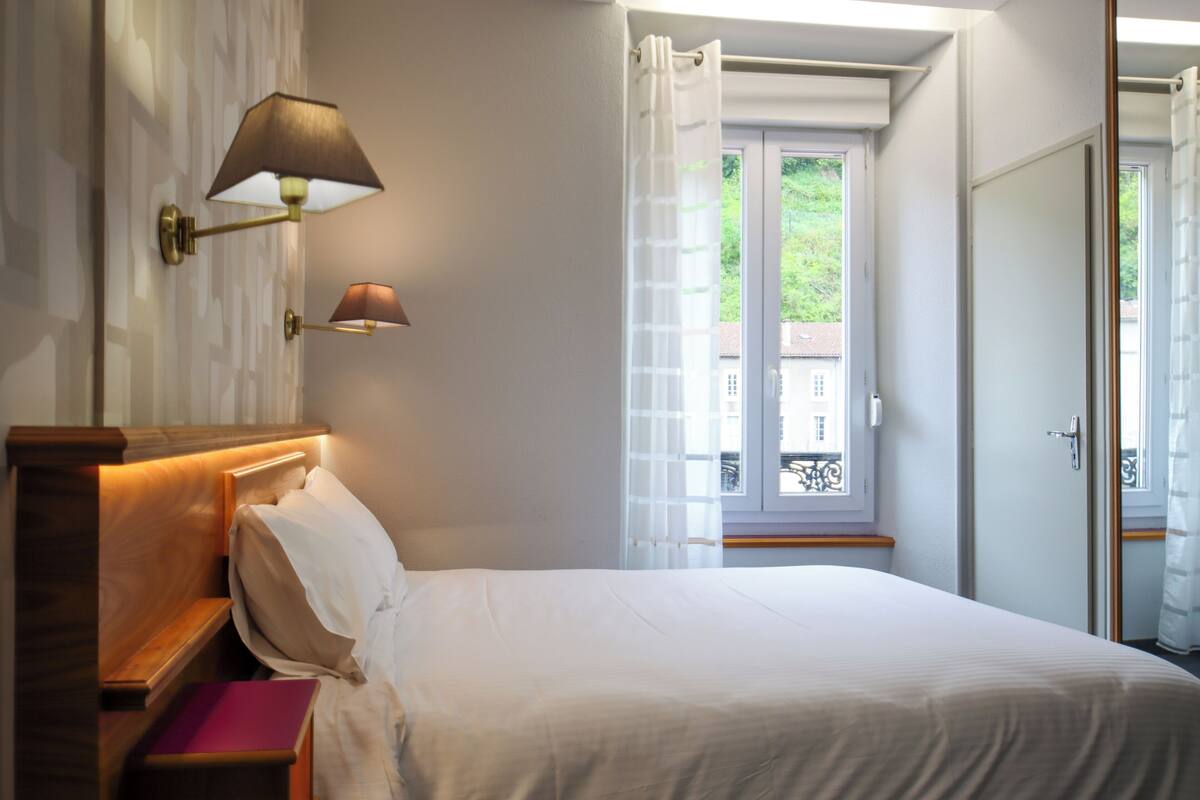 Un dormitorio con cama, mesita de noche, lámpara, ventana con cortinas y puerta.