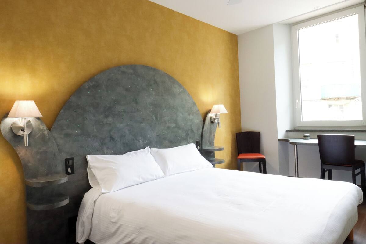 Una habitación de hotel con una cama grande, dos lámparas de noche, un escritorio con una silla y una ventana.