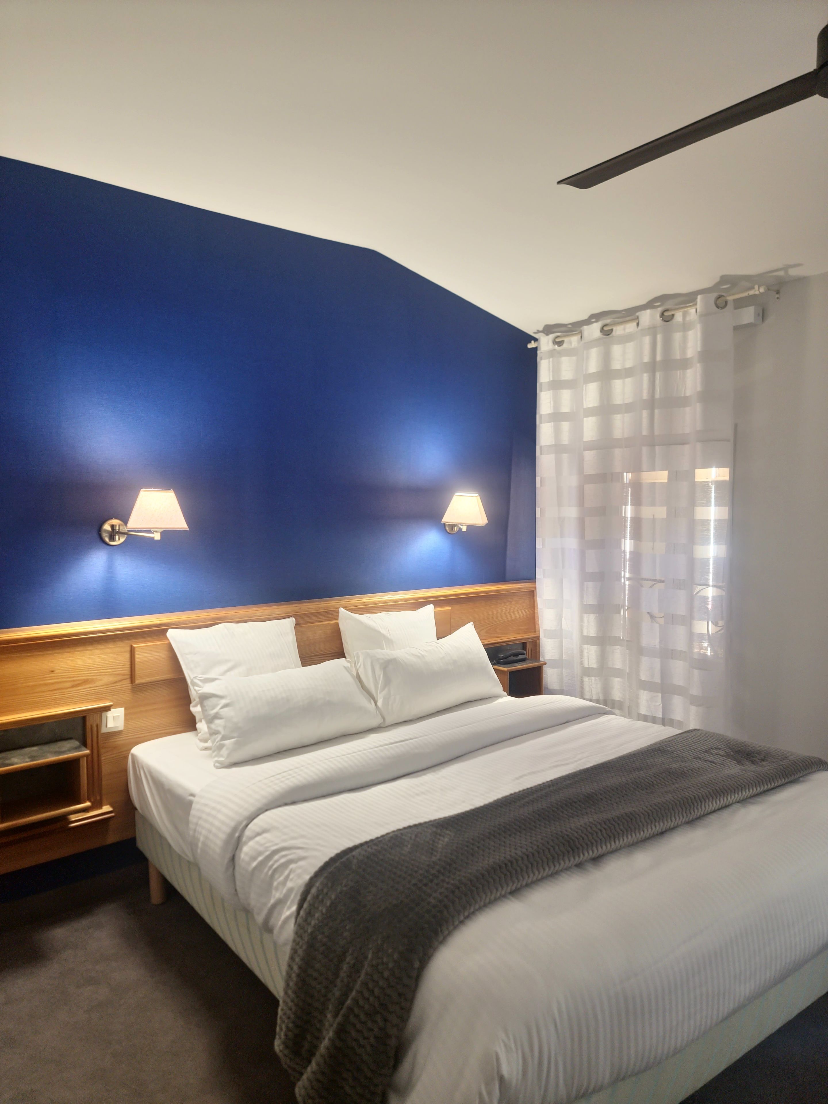 A hotel room with a bed, two bedside lamps, a wooden headboard, a nightstand, and a window with curtains.