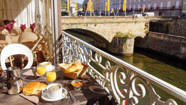 Une table dressée avec des aliments pour le petit-déjeuner, notamment des croissants, du jus d’orange et du café, donnant sur une rivière avec un pont de pierre et des bâtiments en arrière-plan.