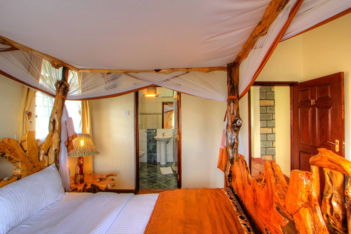Une chambre comprenant un lit, une tête de lit en bois, une table de chevet, une lampe et une porte donnant sur une salle de bain.