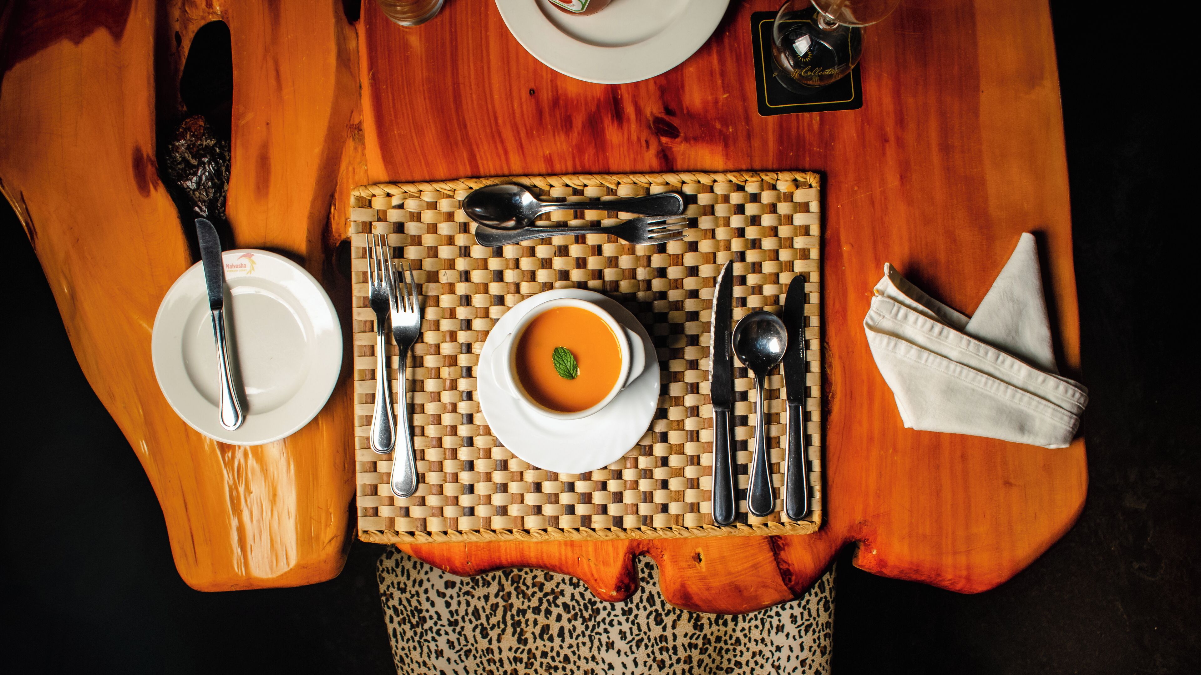 Une table en bois, sur laquelle on a posé un set de table en fibres tressées, un bol de soupe, des couverts et une serviette.
