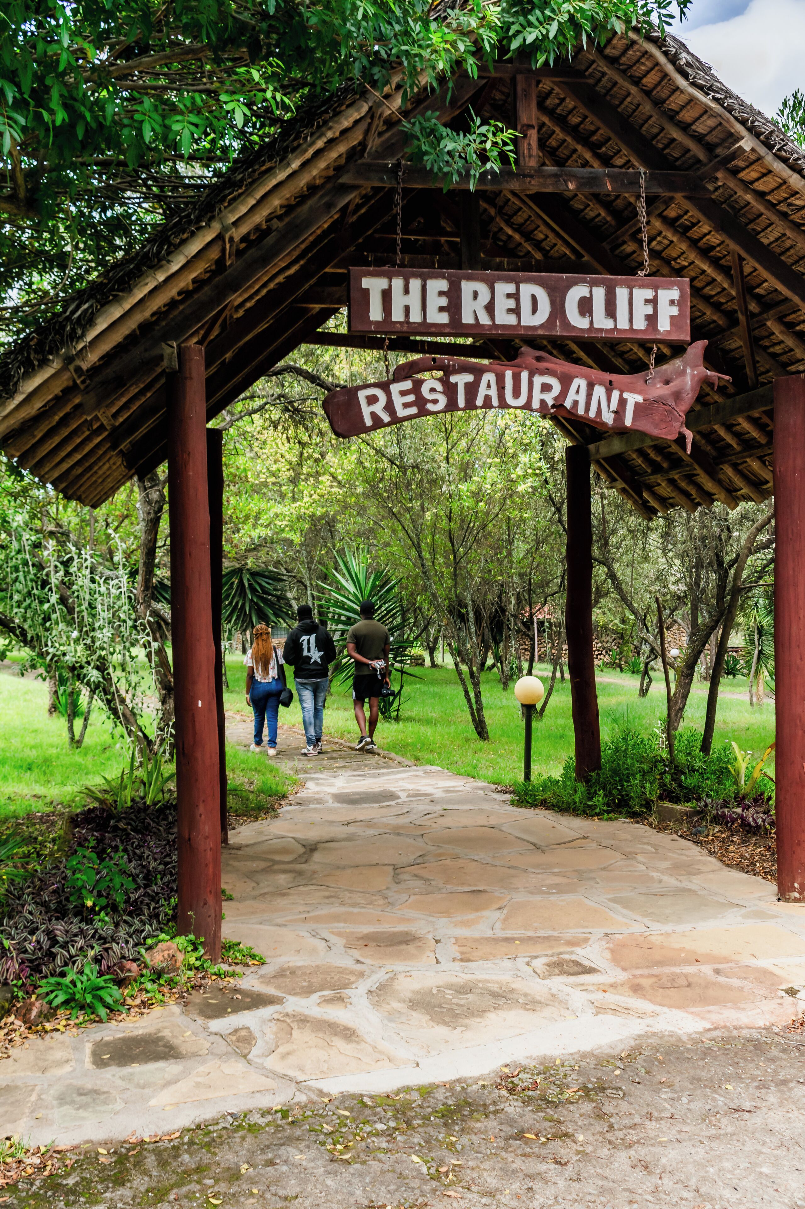 A pathway leading to The Red Cliff Restaurant, surrounded by lush greenery and a wooden structure.