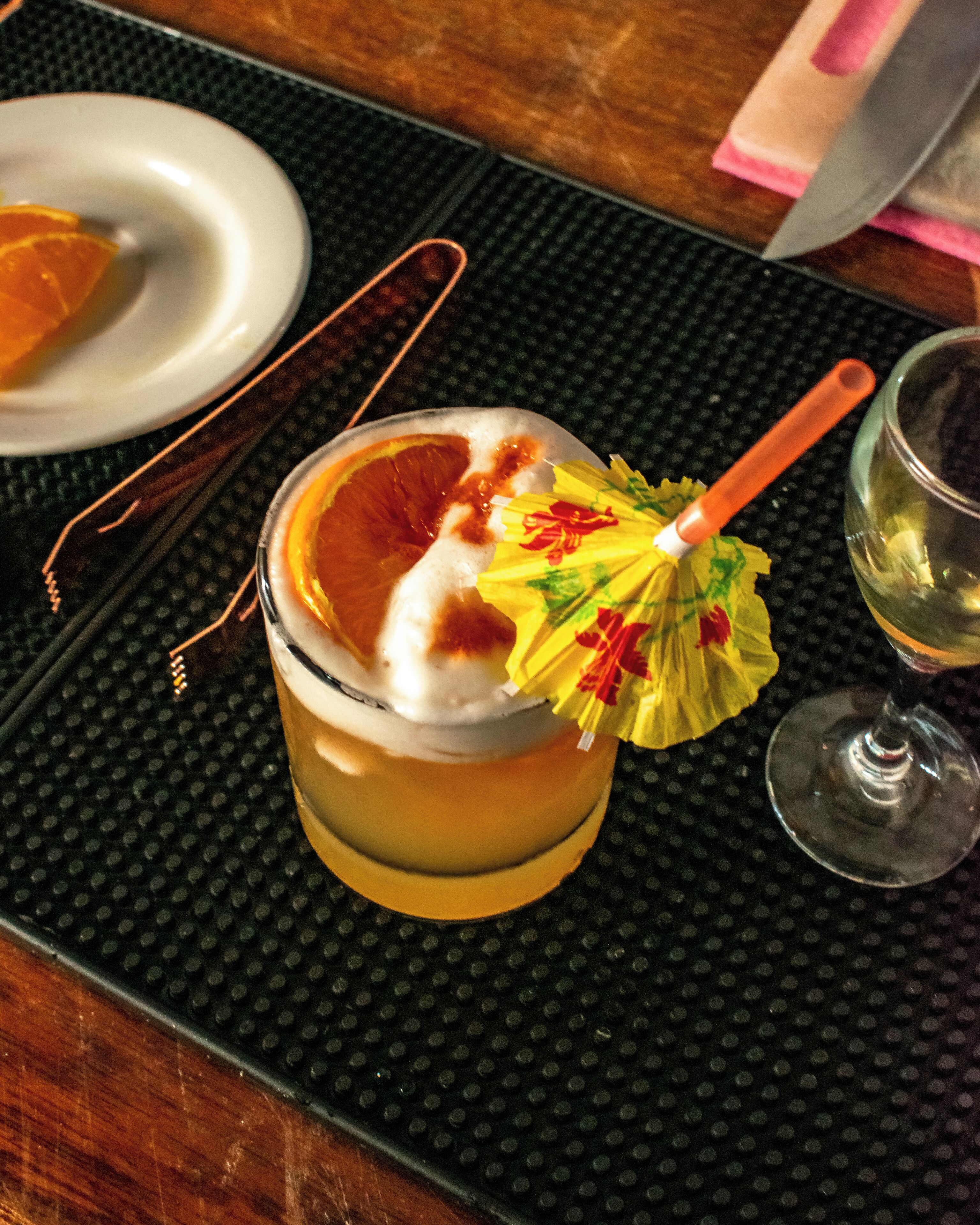 Ein Cocktail mit Orangenscheibe und einer dekorativen Schirmchen auf einem schwarzen, strukturierten Tisch.