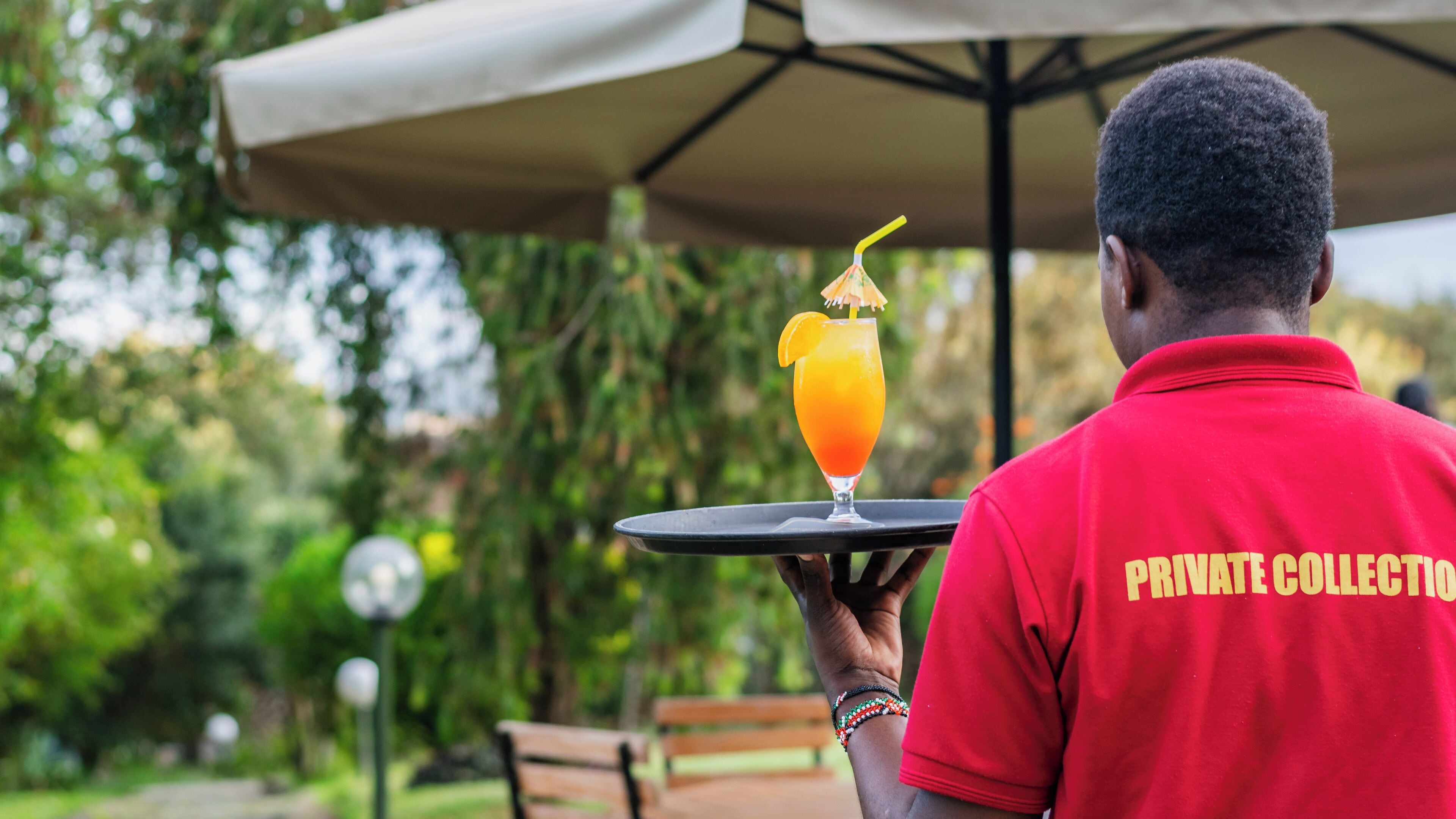 A person in a red shirt with "PRIVATE COLLECTION" text, holding a tray with a drink and a lemon slice.