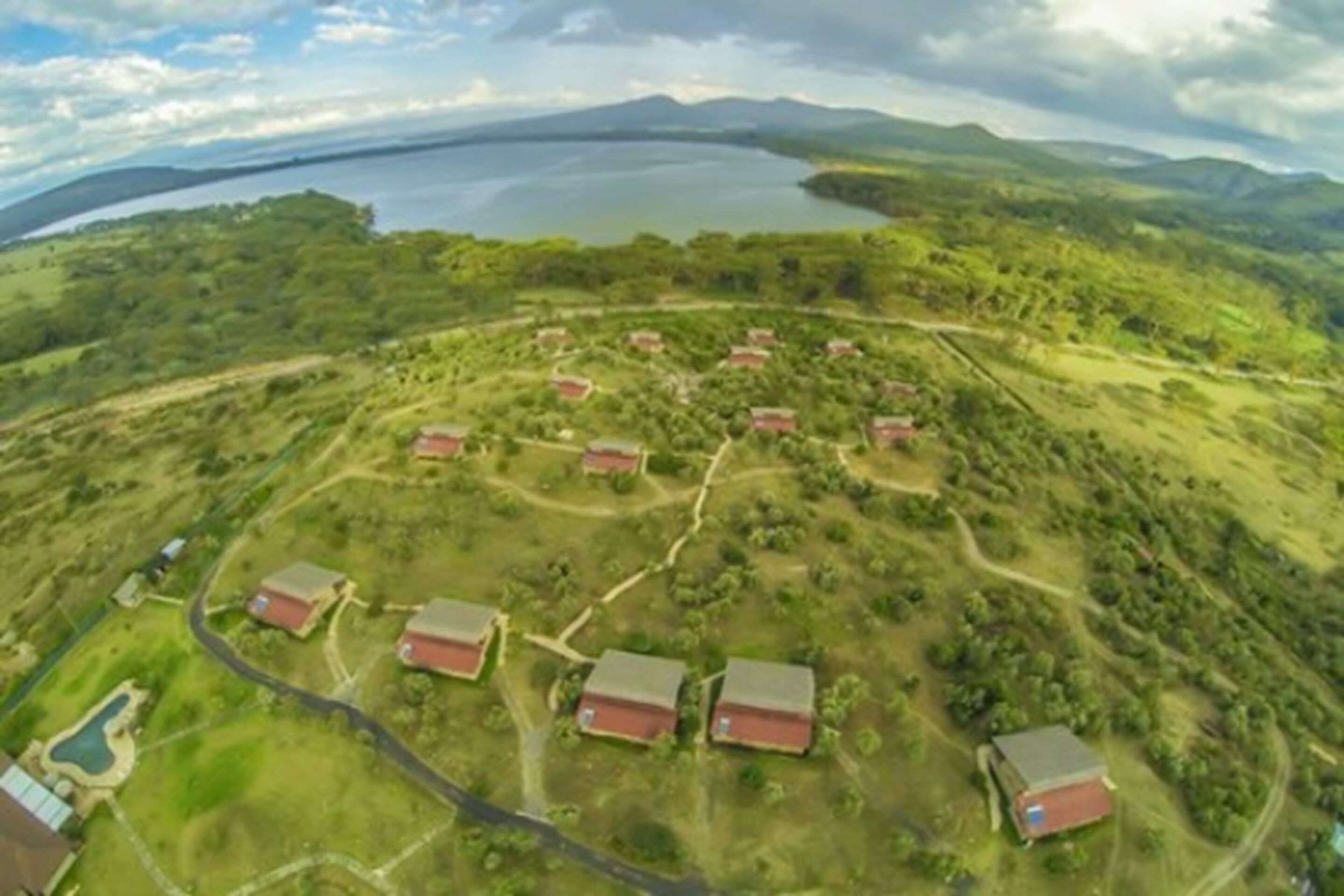 An aerial view of a rural area with several red-roofed buildings arranged in a semi-circular pattern, surrounded by green fields and a large body of water in the background.