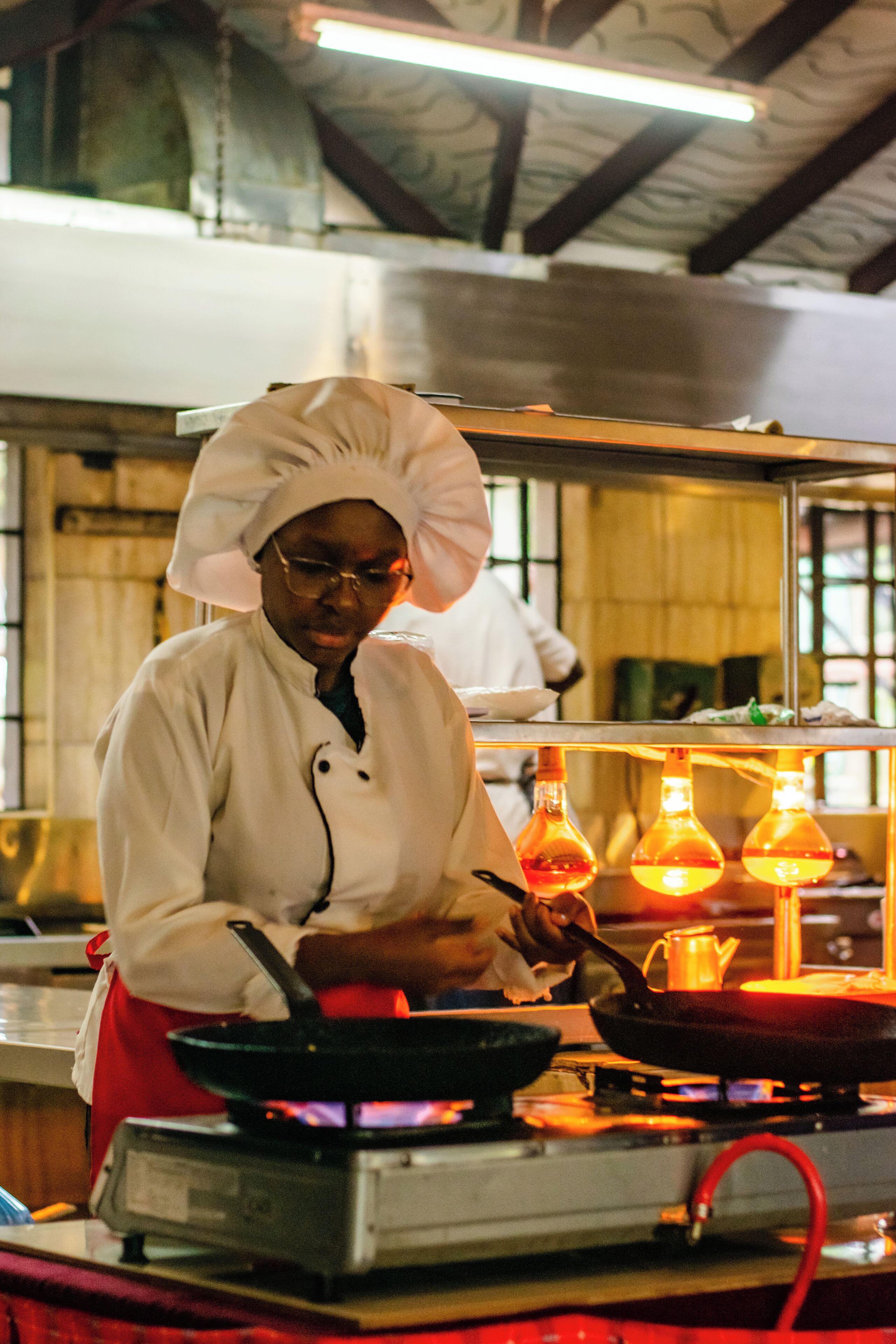 Un chef, vêtu d’une veste et d’un chapeau blancs, cuisine à l’aide d’une poêle sur un réchaud portatif.
