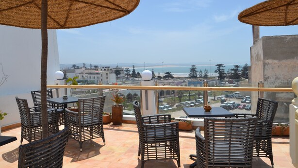 A balcony with wicker furniture, two large woven umbrellas, and a view of the sea and cityscape. - Hotel Riad Benatar (Essaouira)